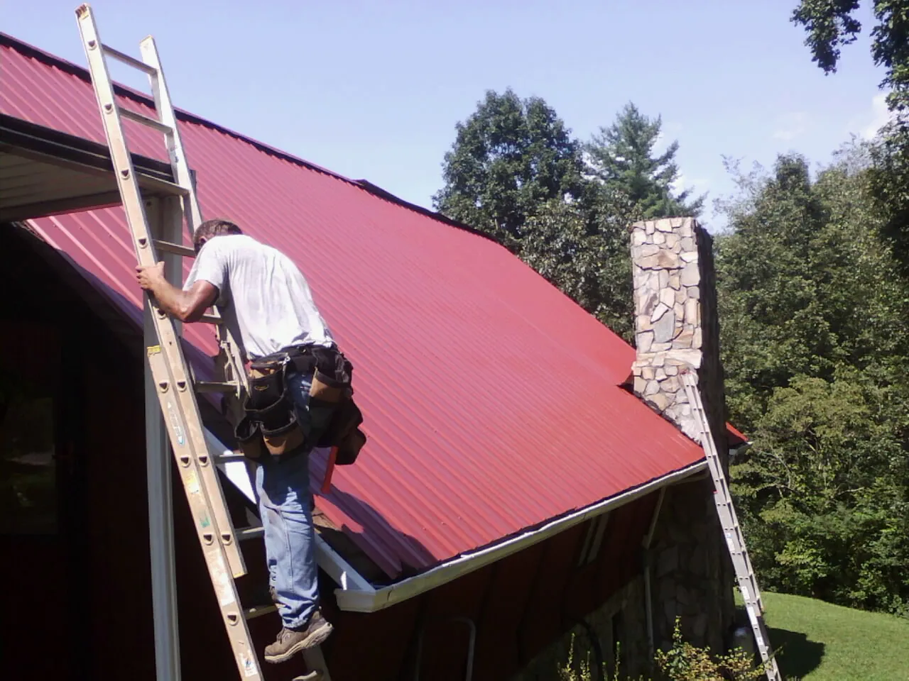 Roofer on a ladder beside a red metal roof and stone chimney — Soffit & Fascia Repair by Roofing Brothers in Katonah
